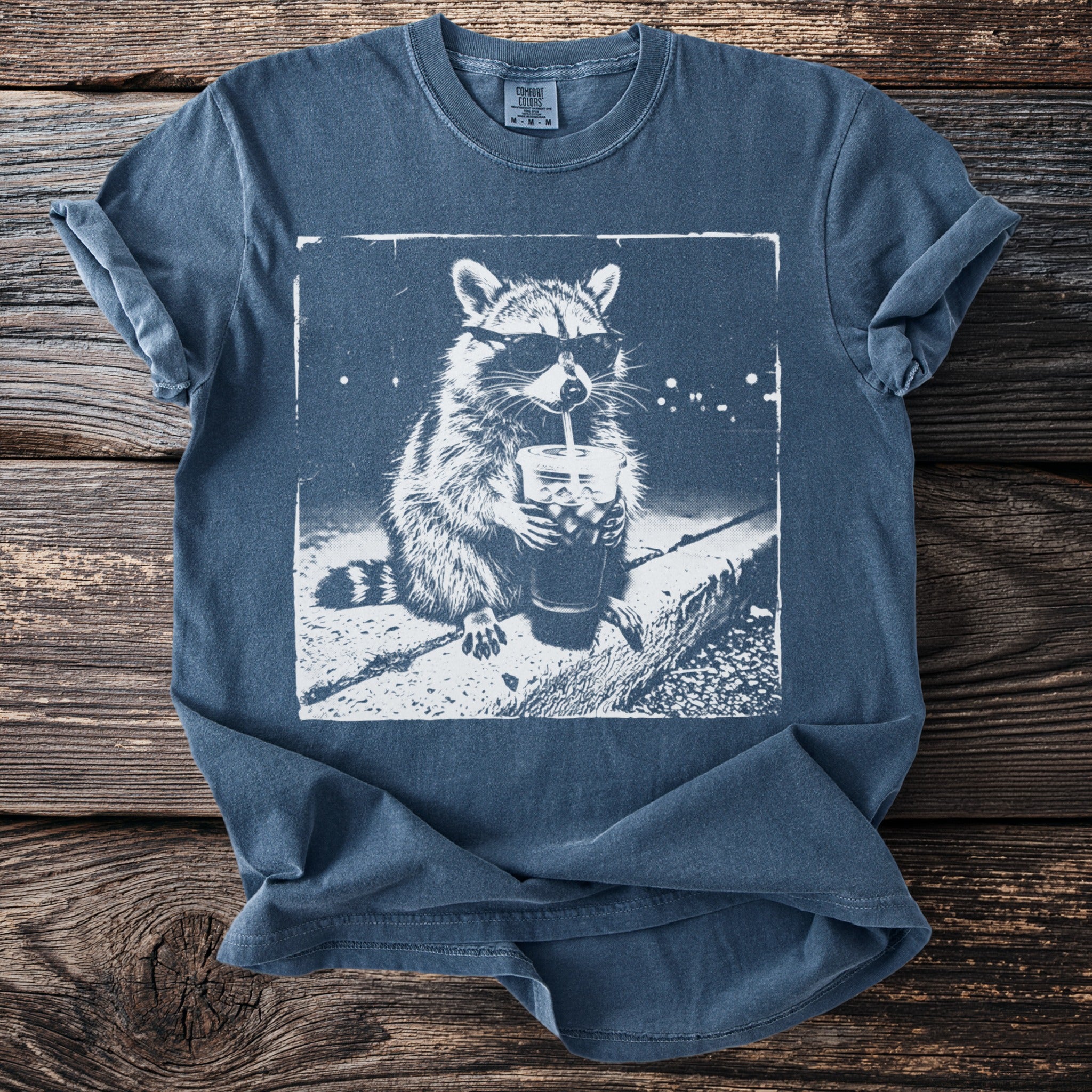 Blue t-shirt with white graphic of a raccoon in sunglasses drinking a beverage.