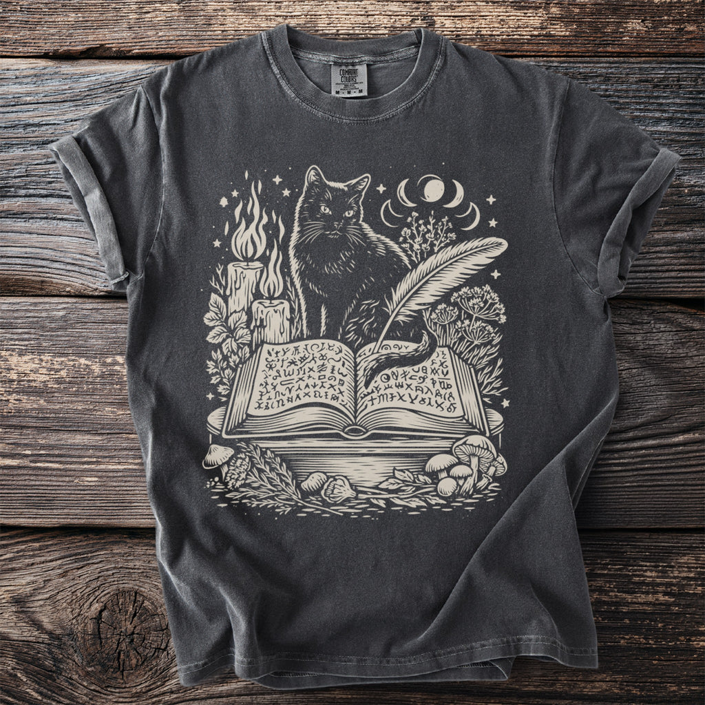 Dark grey t-shirt with the Comfort Colors tag. The graphic features a black cat, an open book, a quill, a candle, and mushrooms.