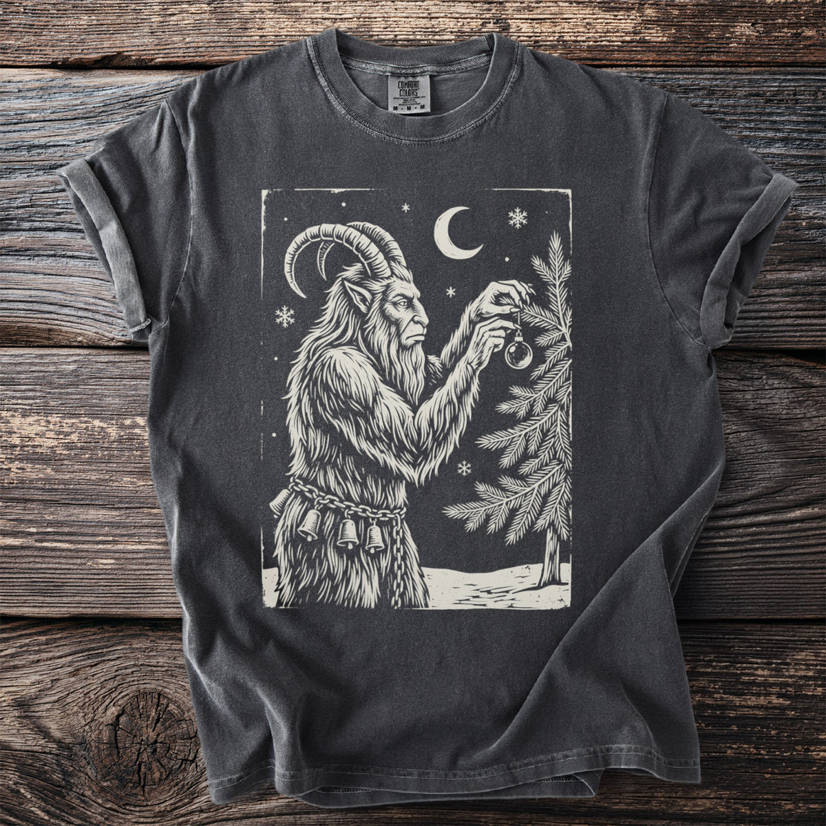 Pepper gray t-shirt with a cream folk-cut print: Krampus hangs one glass ornament on a snowy fir, chains and bells at his waist.