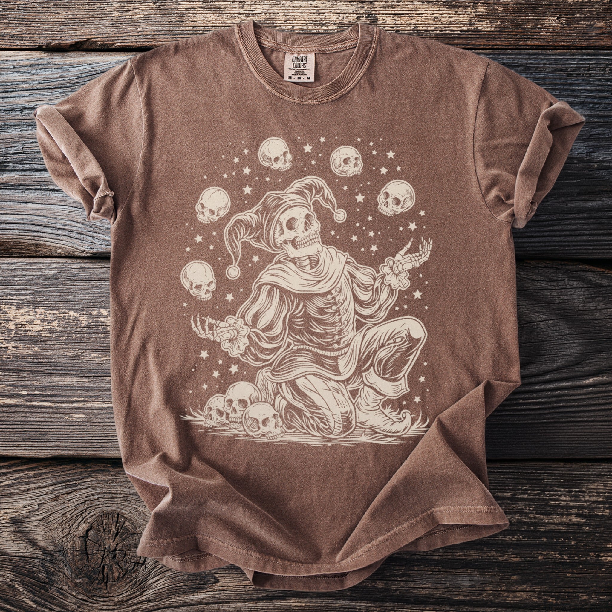 Skeleton jester juggling skulls with stars on a brown t-shirt.