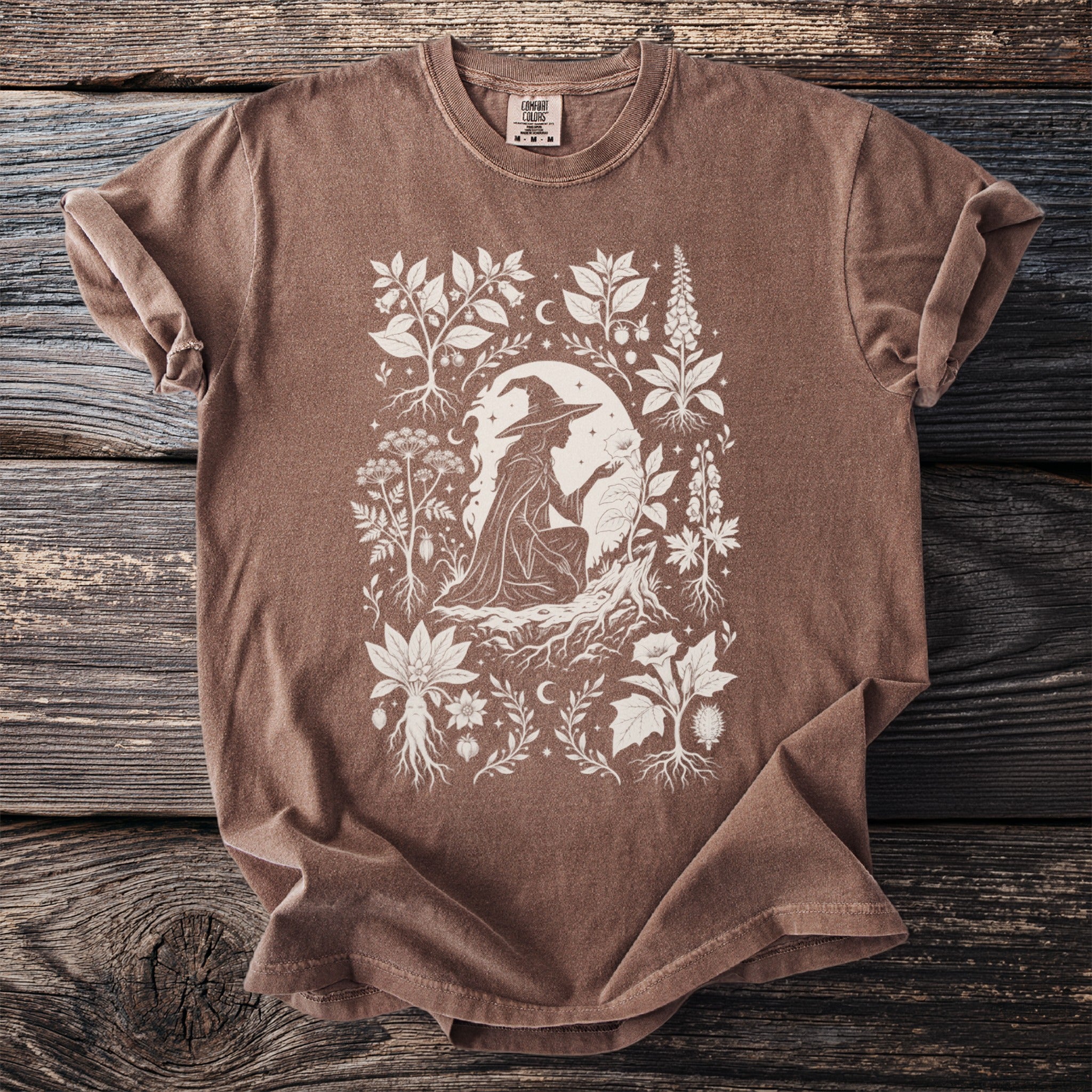 Brown t-shirt with a white line drawing of a witch and botanical elements.