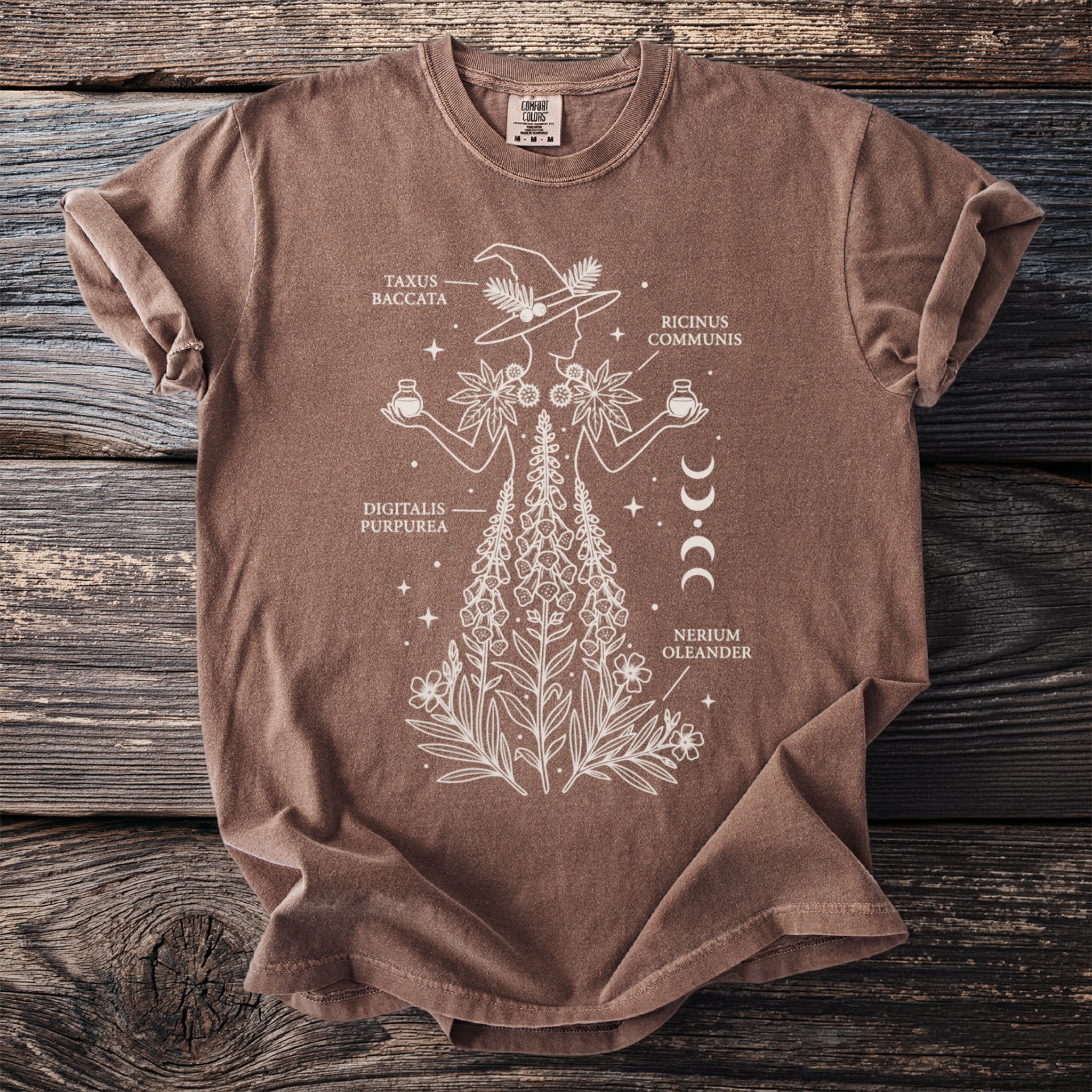Brown t-shirt with white line art of a witch and plants, labeled "Taxus Baccata," "Digitalis Purpurea," and "Ricinus Communis."