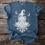 Blue t-shirt with white graphic of a lotus-posed figure wearing a witch hat, moons, stars, and botanical labels.