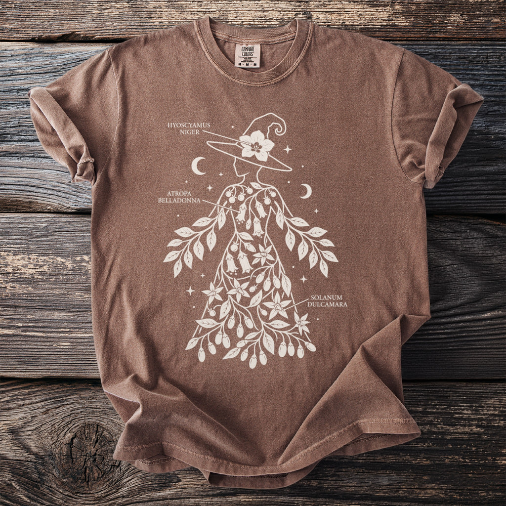 Brown t-shirt with a white botanical illustration of a witch adorned with plants and labeled text.