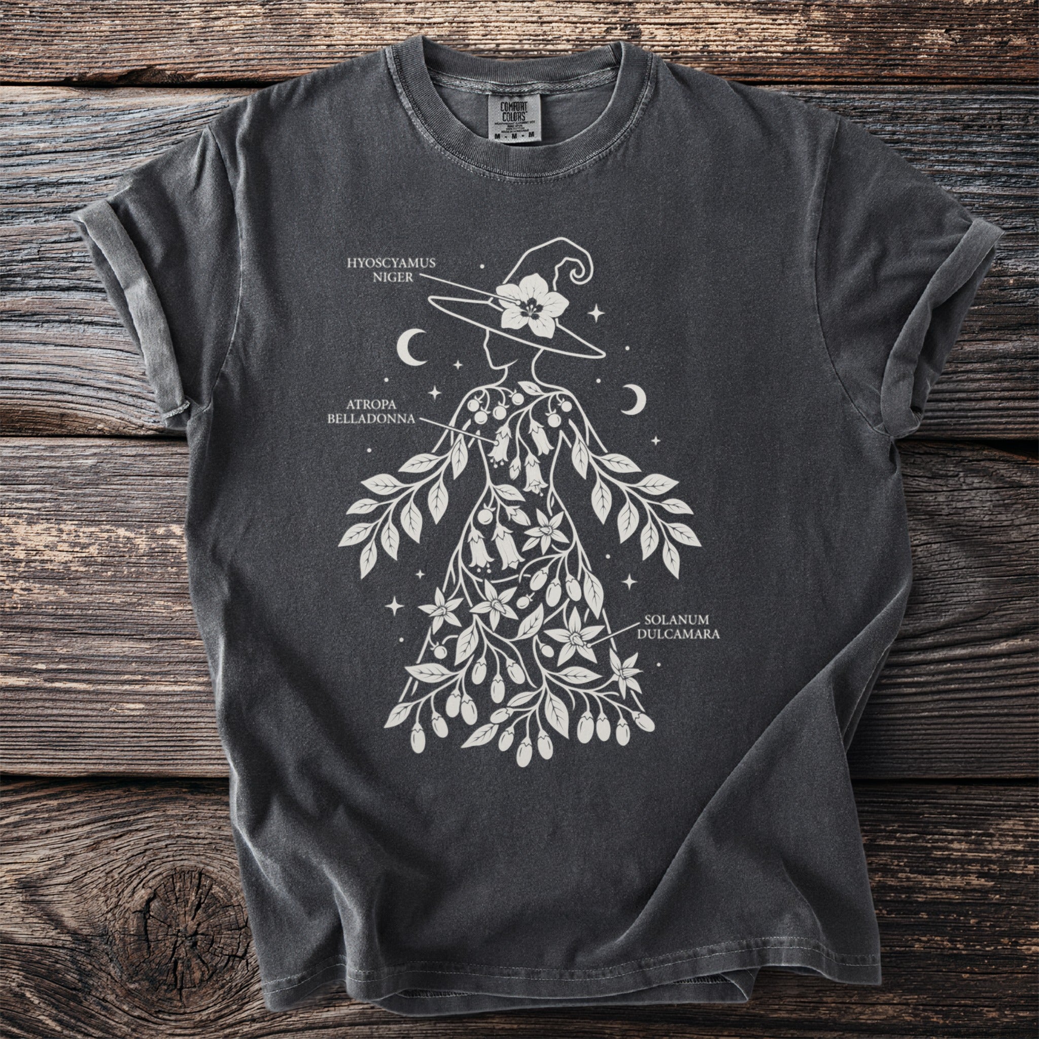 Dark t-shirt features a white outline of a witch adorned with foliage labeled Hyoscyamus Niger, Atropa Belladonna, Solanum Dulcamara.