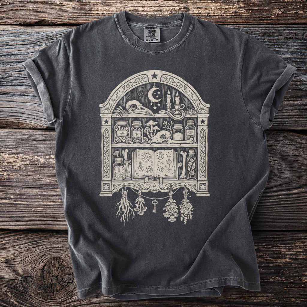Dark gray t-shirt with a pagan-themed graphic of a shelf containing skulls, jars, and a spellbook.