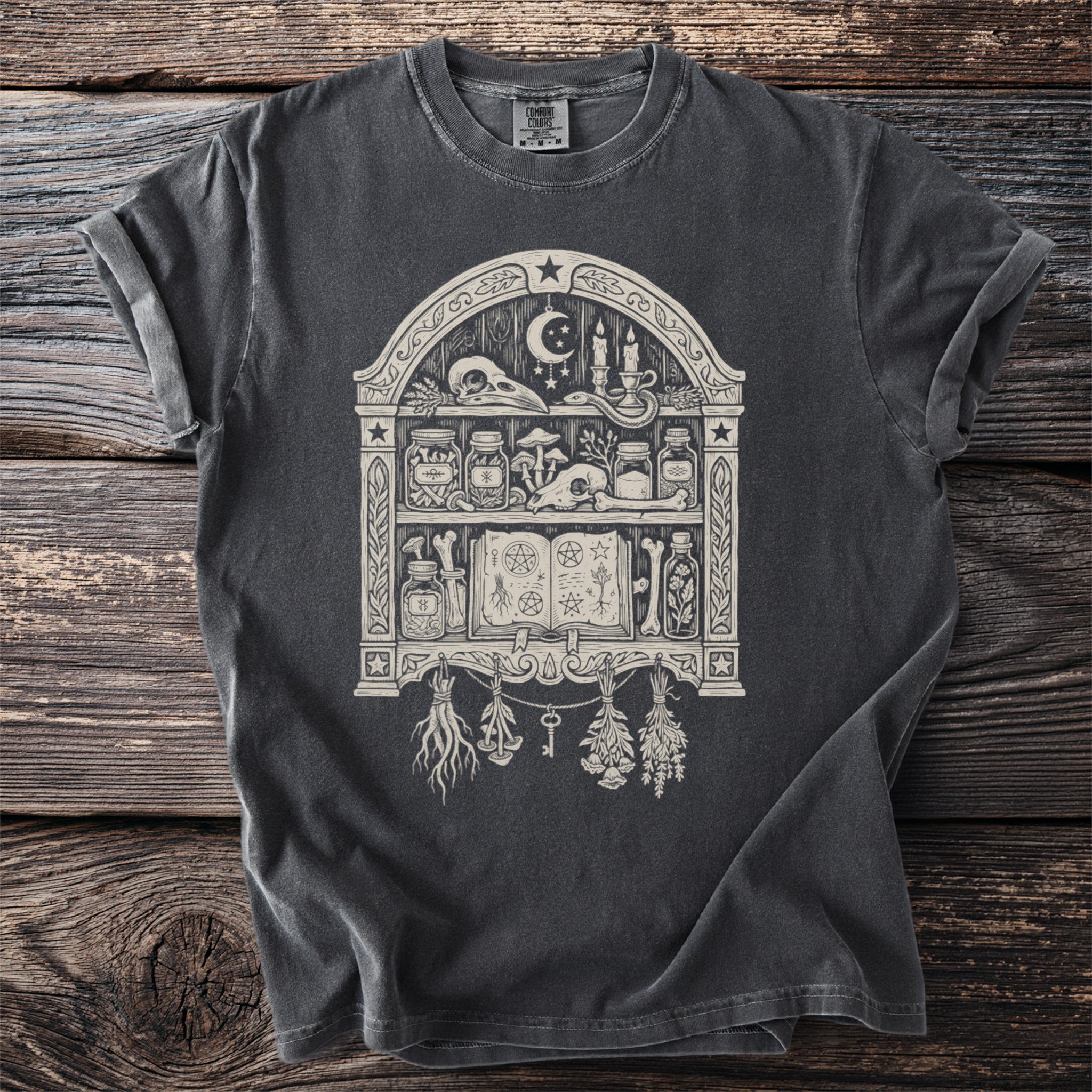 Dark gray t-shirt with a pagan-themed graphic of a shelf containing skulls, jars, and a spellbook.