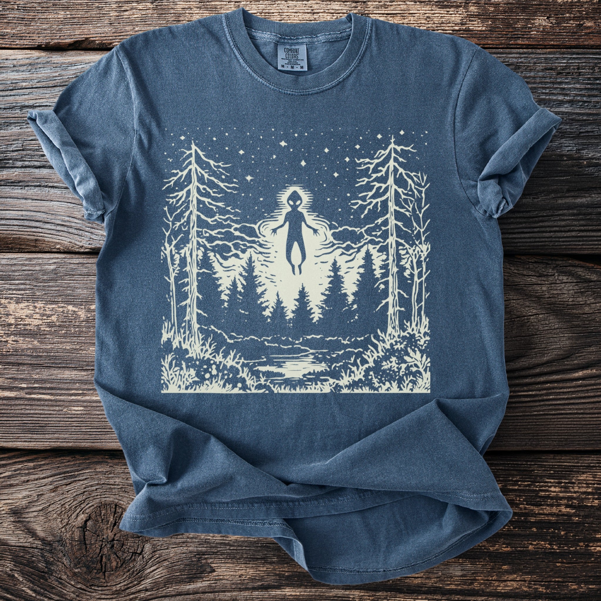 Blue t-shirt with a white graphic of an alien figure floating over a forest at night.