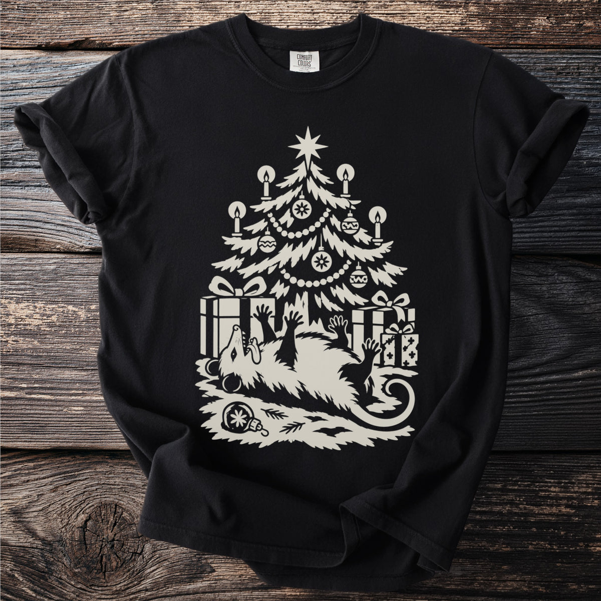 Black t-shirt with a cream folk-cut print: an opossum lies on its back under a candlelit Christmas tree, playing dead among the presents.