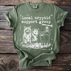 Green t-shirt with "local cryptid support group" text and raccoon, opossum, frog, bat, cat graphic.