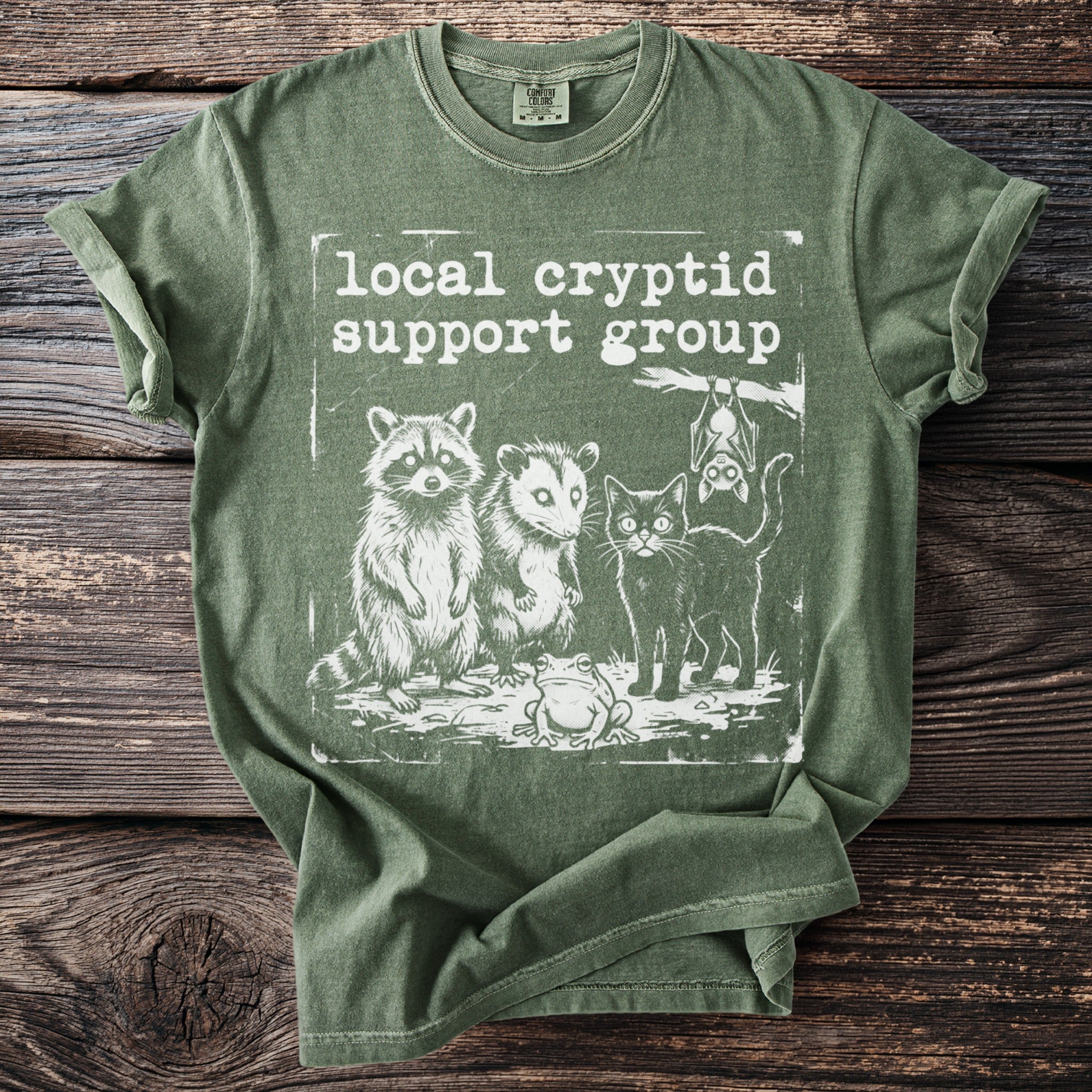 Green t-shirt with "local cryptid support group" text and raccoon, opossum, frog, bat, cat graphic.