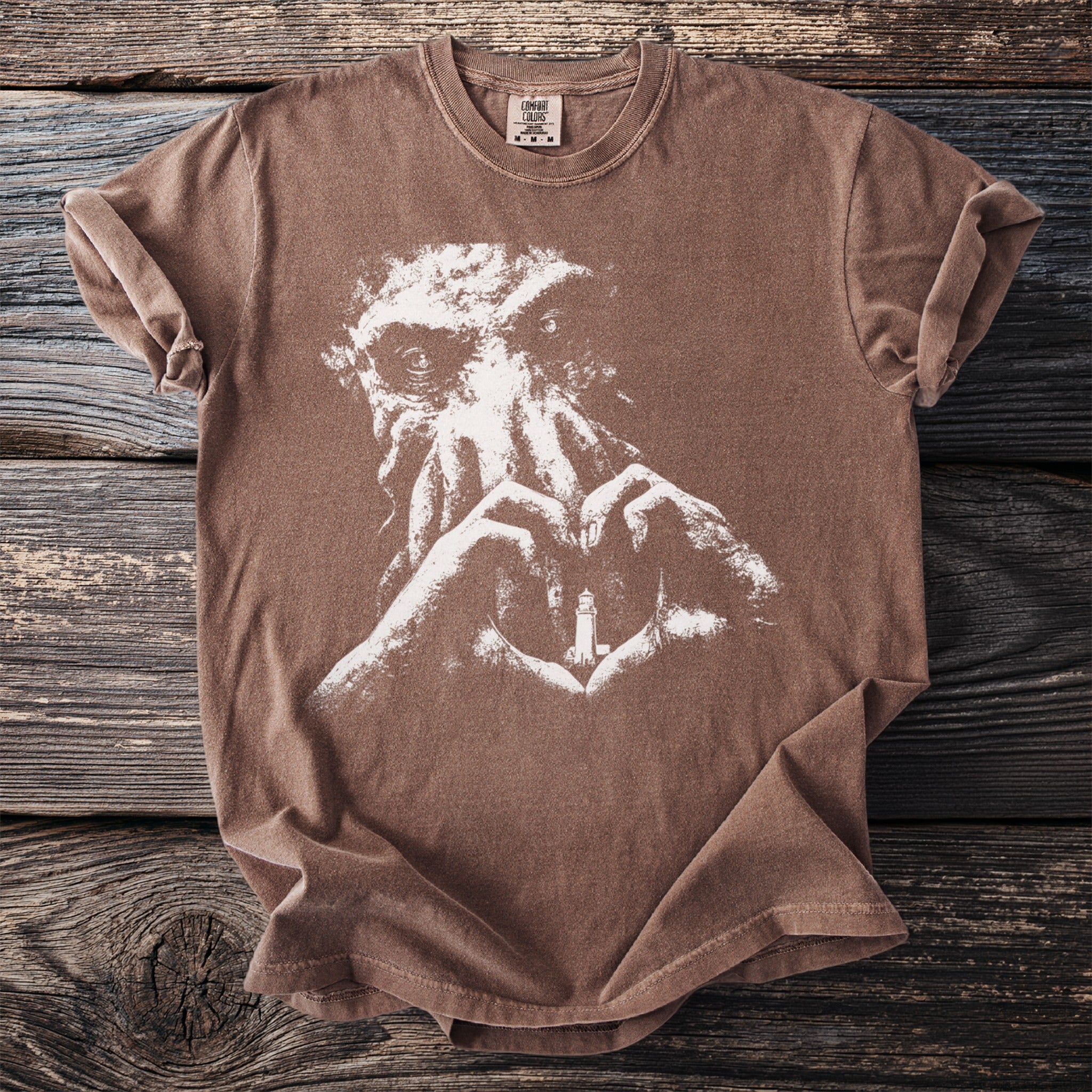Brown t-shirt with white graphic of a creature and hands forming a heart with a lighthouse.