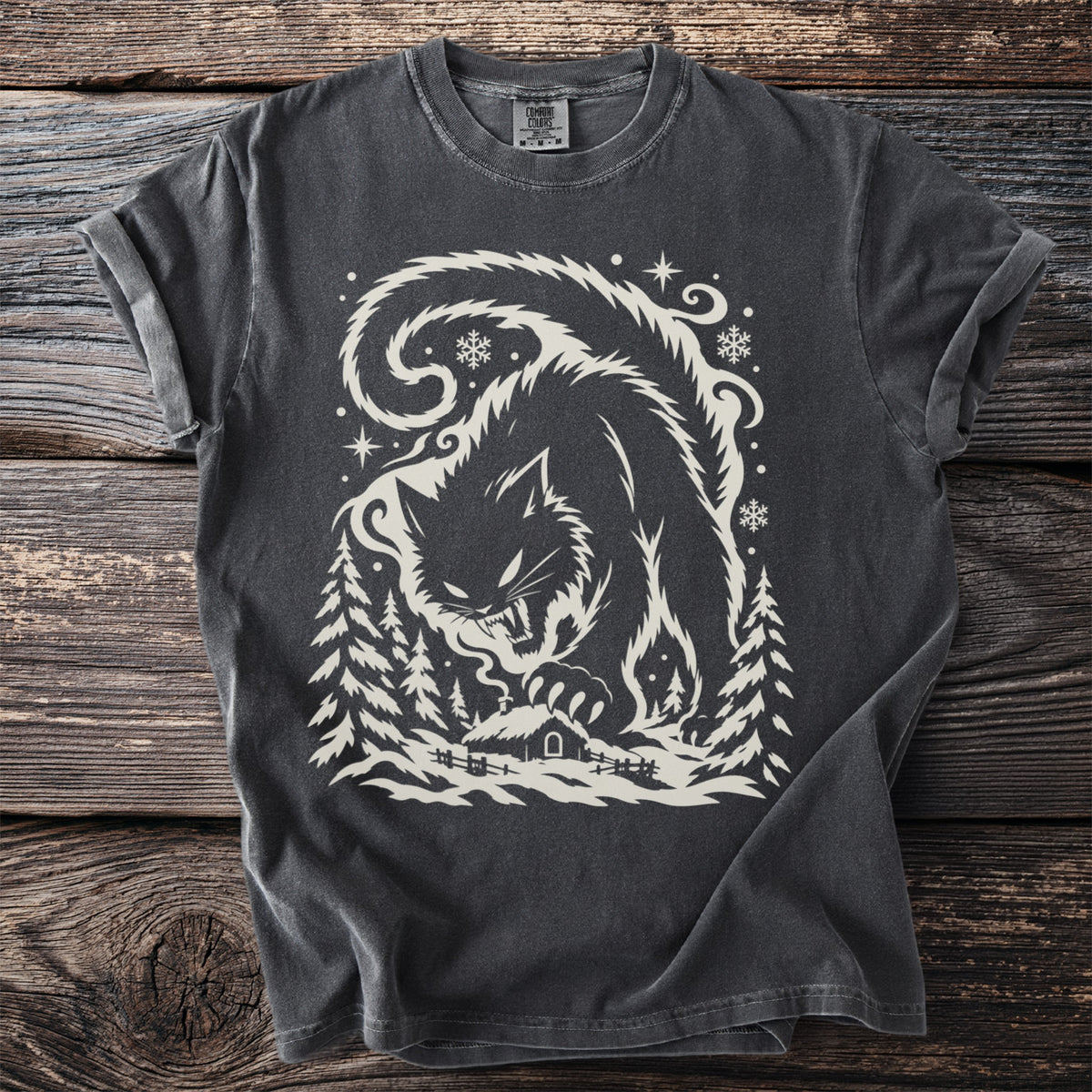 Pepper gray t-shirt with a cream folk-cut print: the enormous Yule Cat, back arched and fangs bared, rests a paw on a small snowy turf cabin among pine trees and snowflakes.
