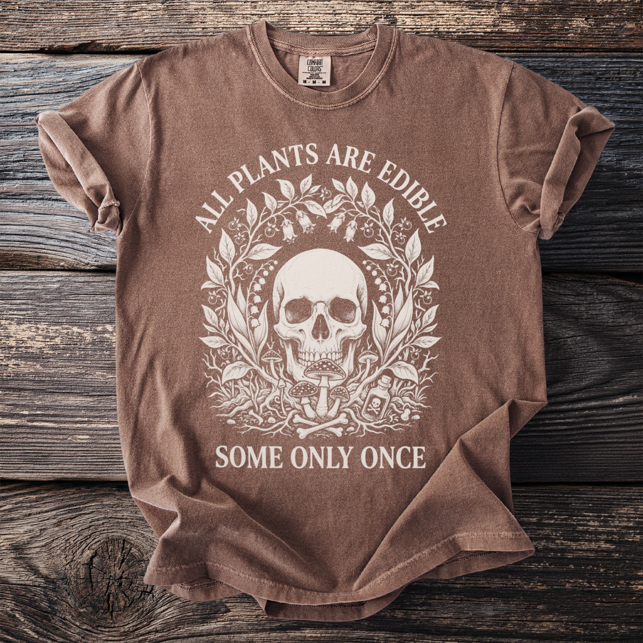 Brown t-shirt with a skull, plants, mushrooms, and bones graphic and text.