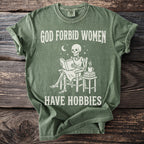 Green t-shirt with skeleton reading a book on chair, "GOD FORBID WOMEN HAVE HOBBIES".