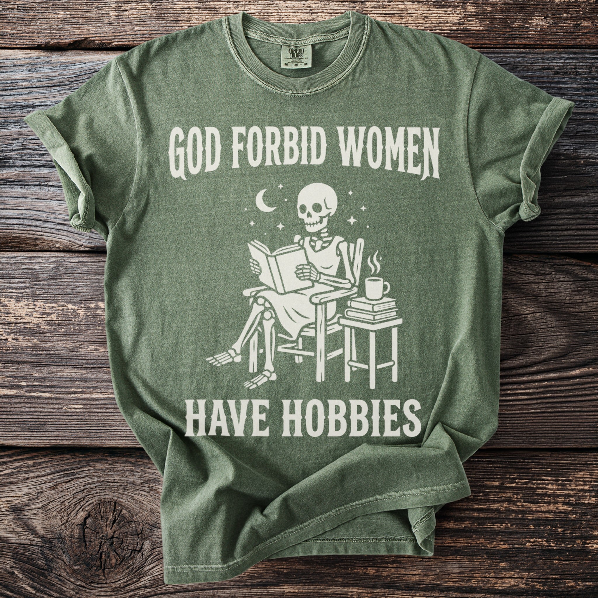 Green t-shirt with skeleton reading a book on chair, "GOD FORBID WOMEN HAVE HOBBIES".