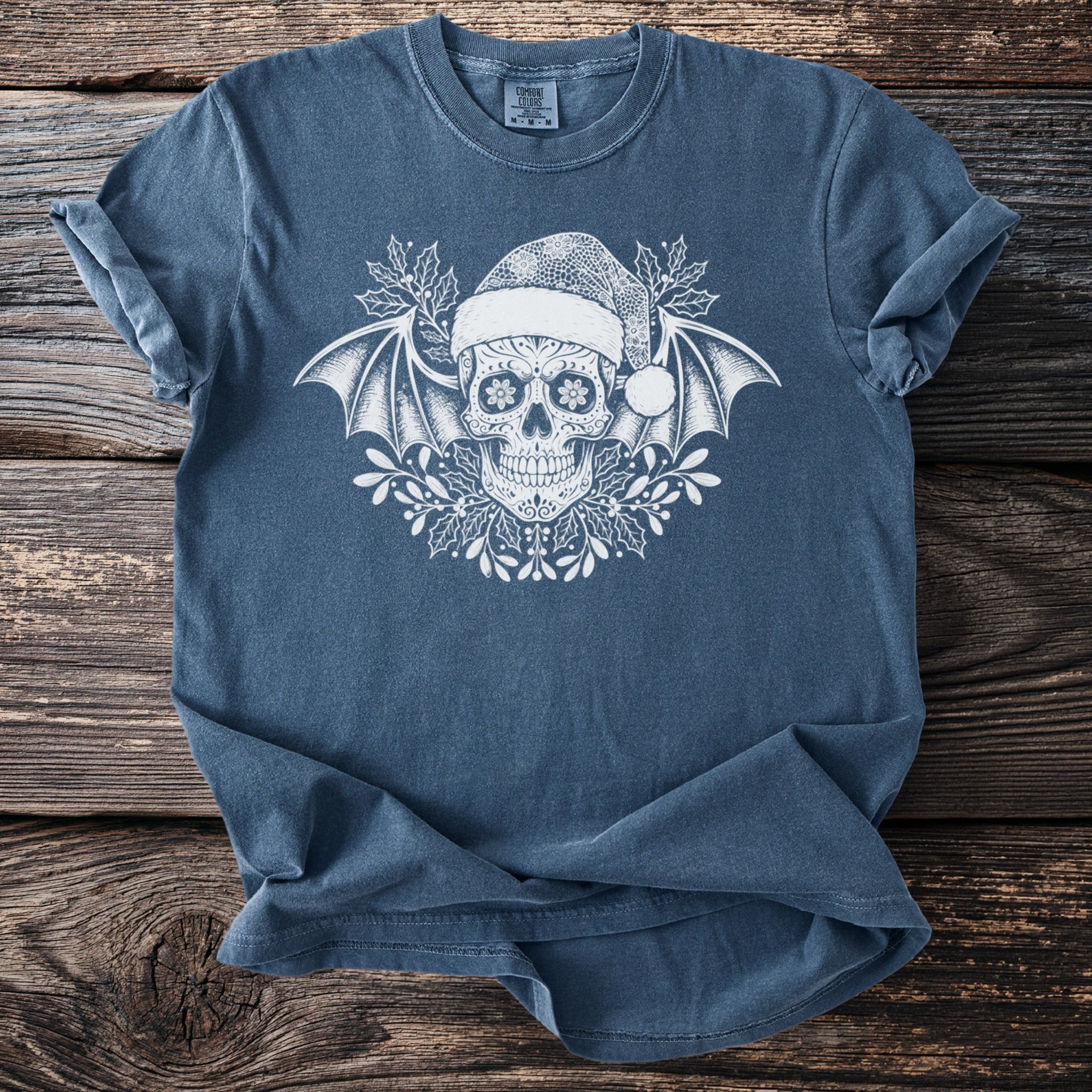 Blue t-shirt with white sugar skull wearing Santa hat, bat wings, and holly design.