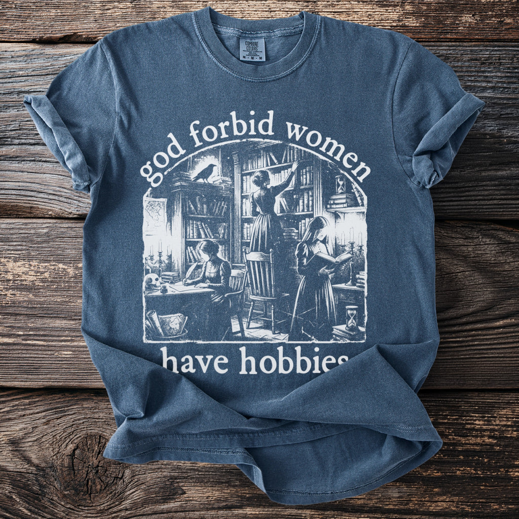 Blue t-shirt with graphic "god forbid women have hobbies" and illustration.