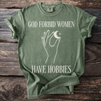 Green t-shirt with white text "GOD FORBID WOMEN HAVE HOBBIES" and a hand holding a moon.