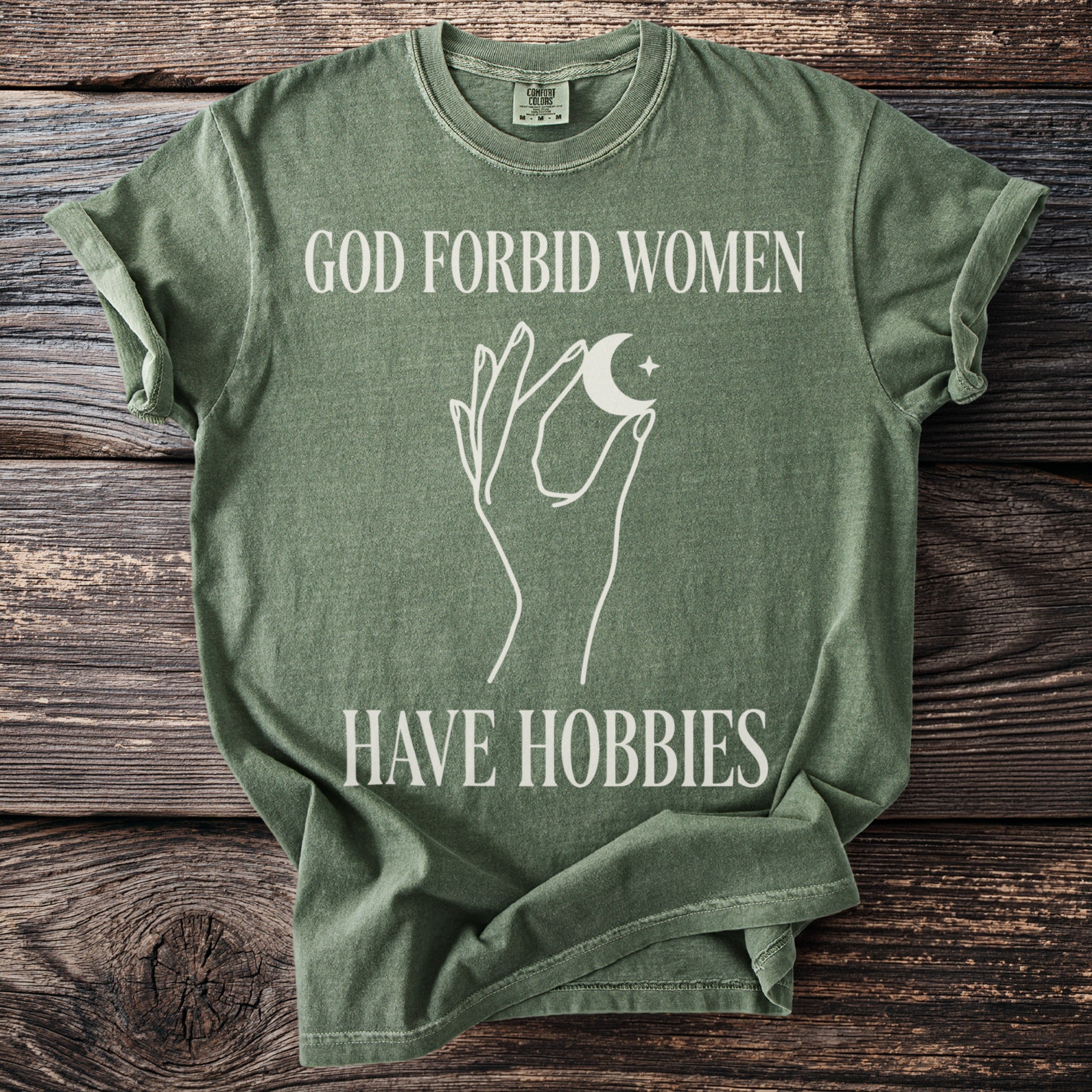 Green t-shirt with white text "GOD FORBID WOMEN HAVE HOBBIES" and a hand holding a moon.
