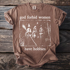 Brown t-shirt with "god forbid women have hobbies" text and image of three witches.