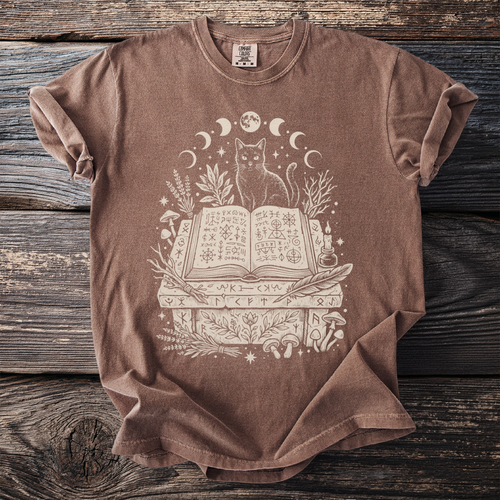Brown t-shirt with a white line drawing of a cat, moon phases, and an open spell book.