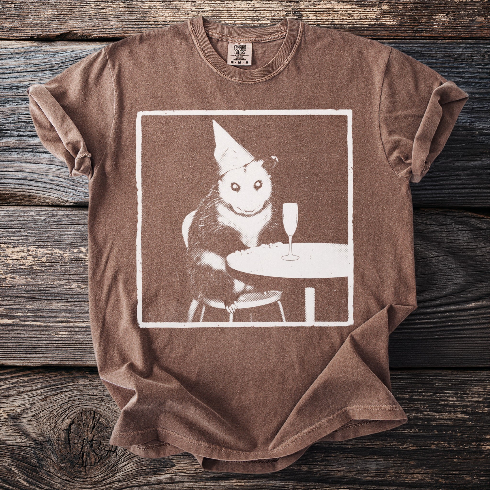 Brown t-shirt with a white graphic of an opossum in a party hat with a champagne flute.