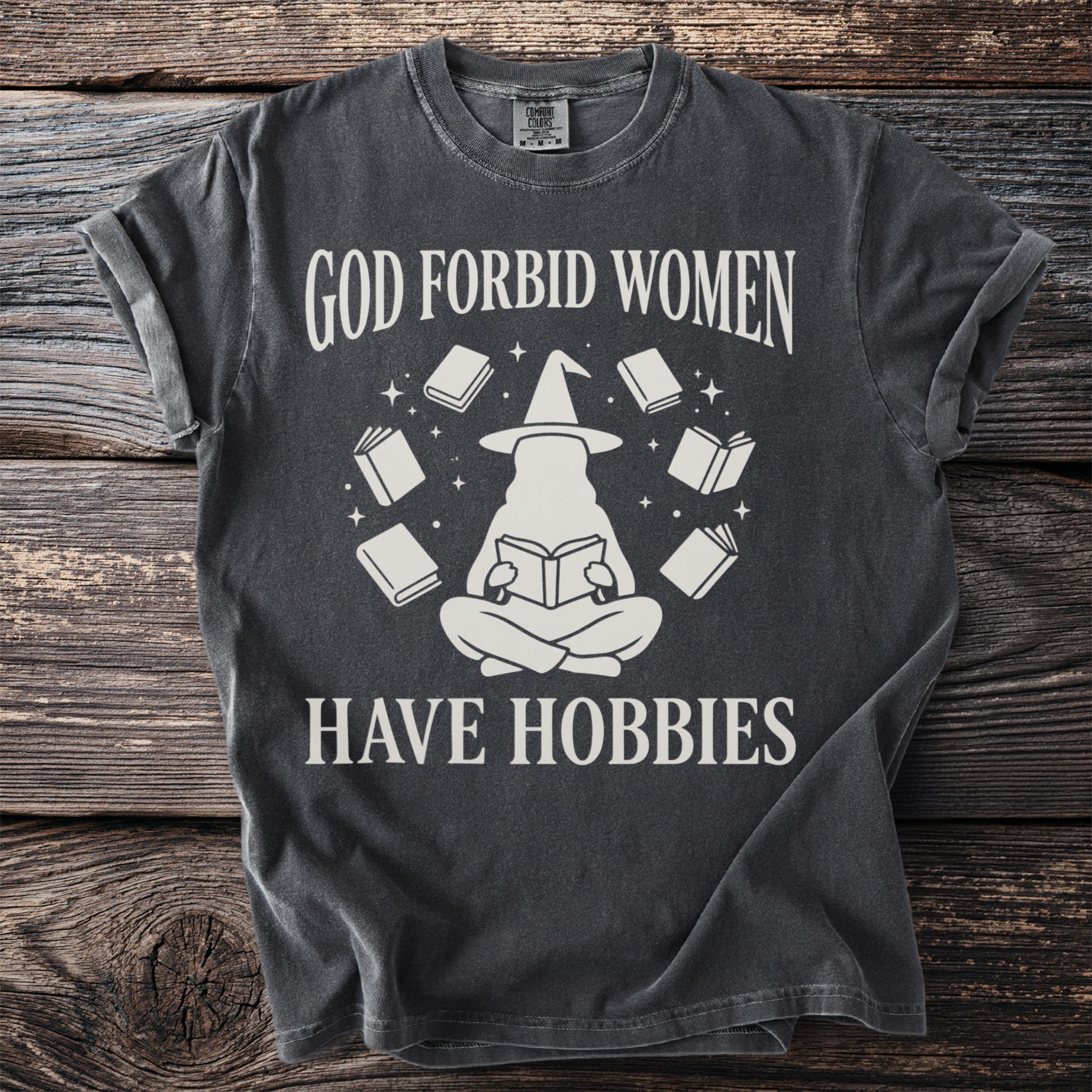 T-shirt with text "God forbid women have hobbies" and a graphic of a witch reading surrounded by books.