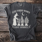 Dark gray t-shirt with text "God Forbid Women Have Hobbies" and a witch watering plants.