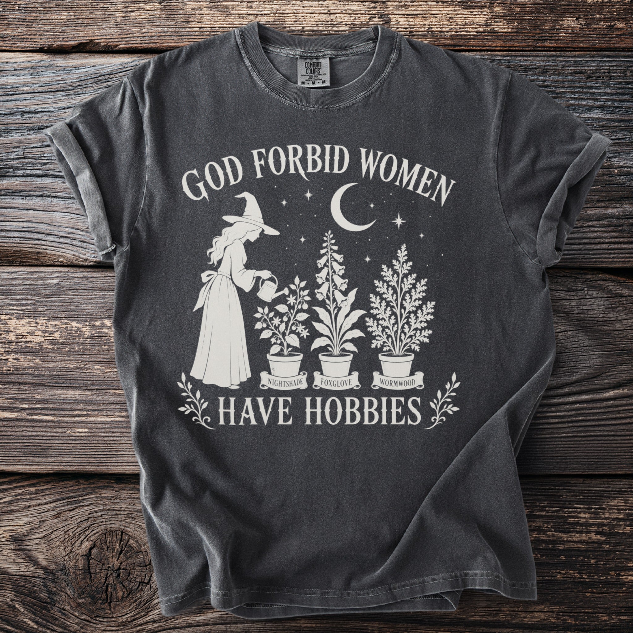 Dark gray t-shirt with text "God Forbid Women Have Hobbies" and a witch watering plants.