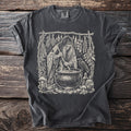 Dark t-shirt with a witch stirring a cauldron surrounded by herbs, mushrooms and a raven.