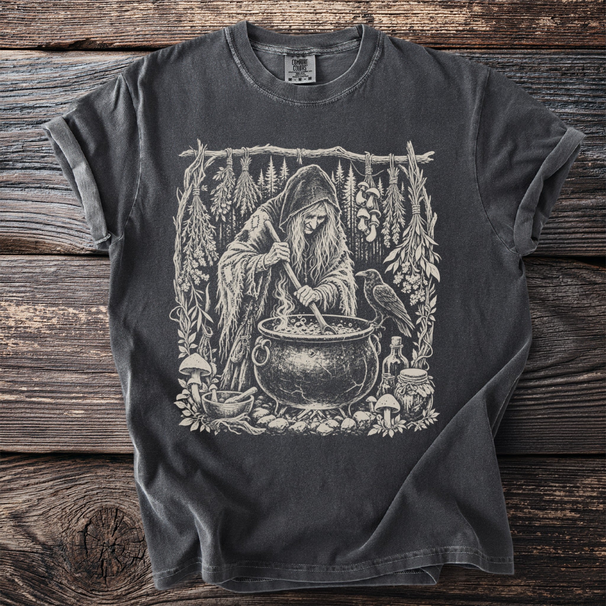 Dark t-shirt with a witch stirring a cauldron surrounded by herbs, mushrooms and a raven.