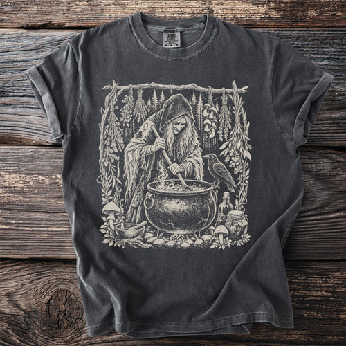 Dark t-shirt with a witch stirring a cauldron surrounded by herbs, mushrooms and a raven.
