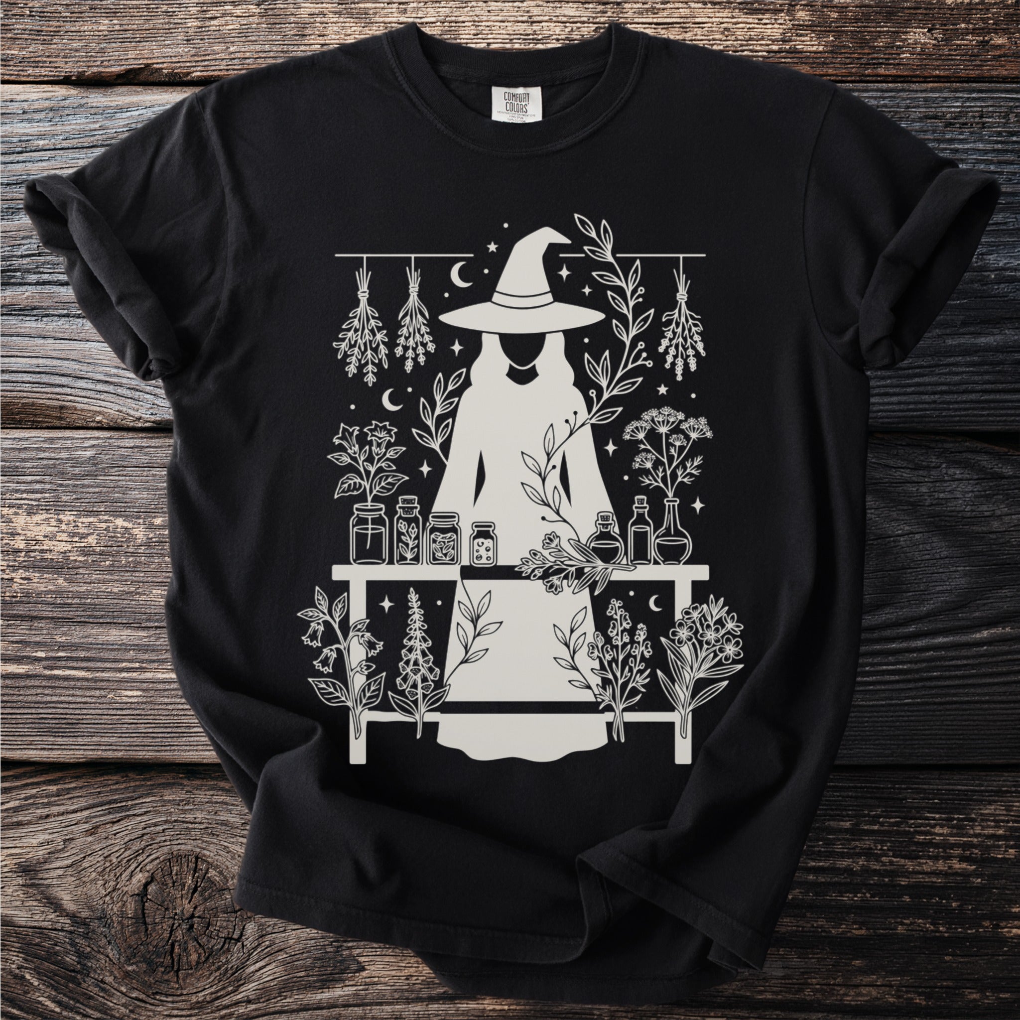 Black t-shirt with white line art of a witch and potion bottles surrounded by herbs.