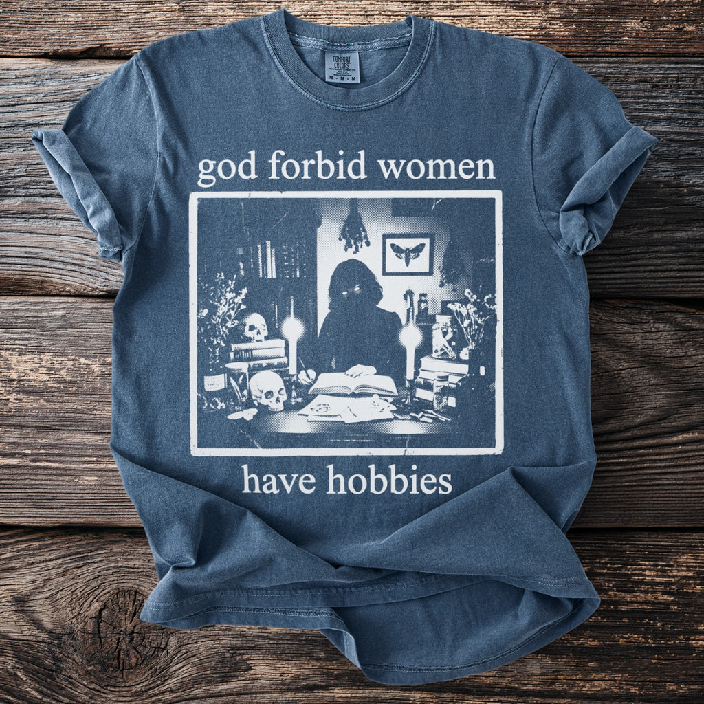Blue t-shirt with "god forbid women have hobbies" and a sketch of a figure at a desk.