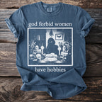 Blue t-shirt with "god forbid women have hobbies" and a sketch of a figure at a desk.