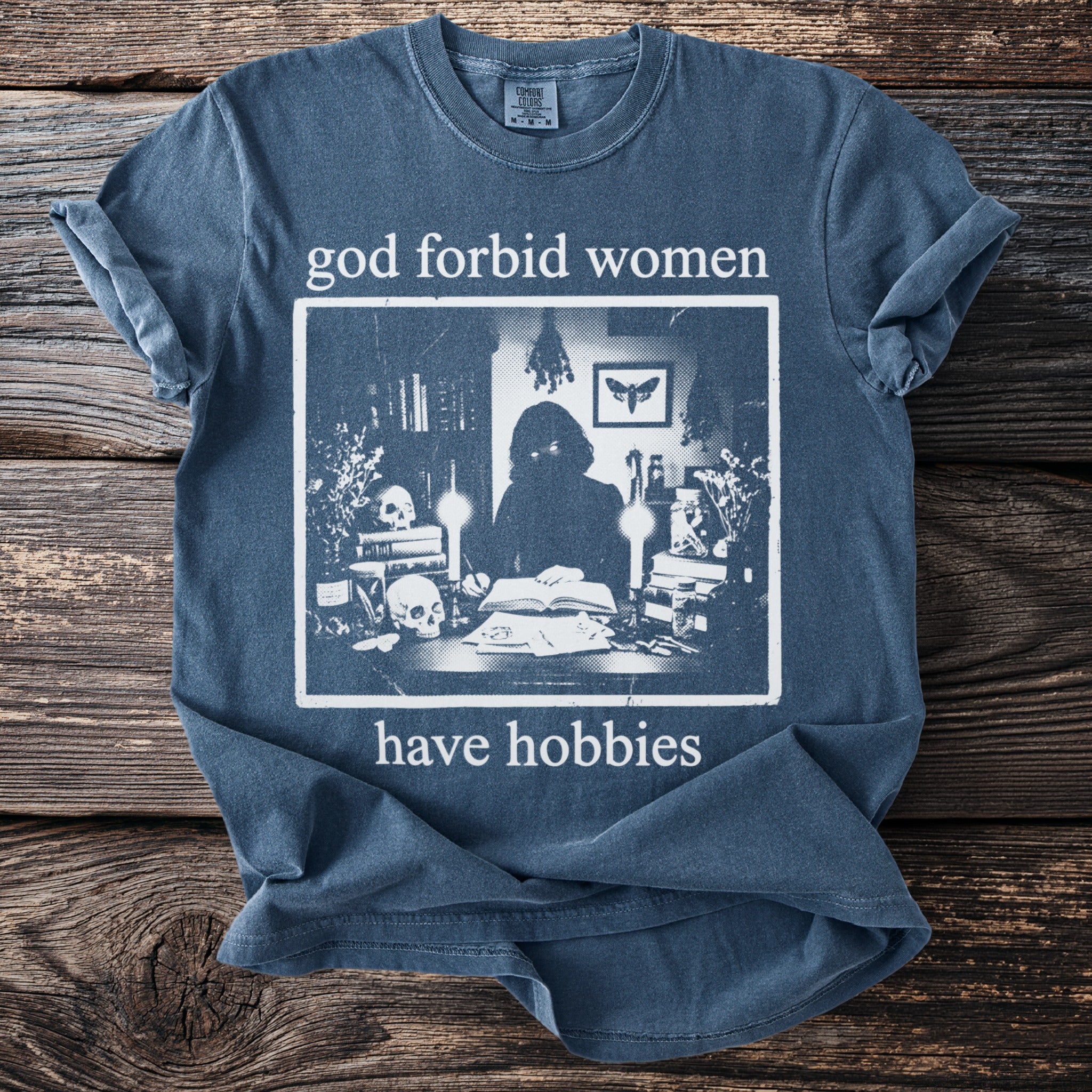 Blue t-shirt with "god forbid women have hobbies" and a sketch of a figure at a desk.