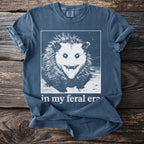 Blue t-shirt with a white graphic of a screaming possum and text "in my feral era".