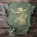 Green t-shirt with a tan graphic of a Highland cow wearing a cowboy hat.