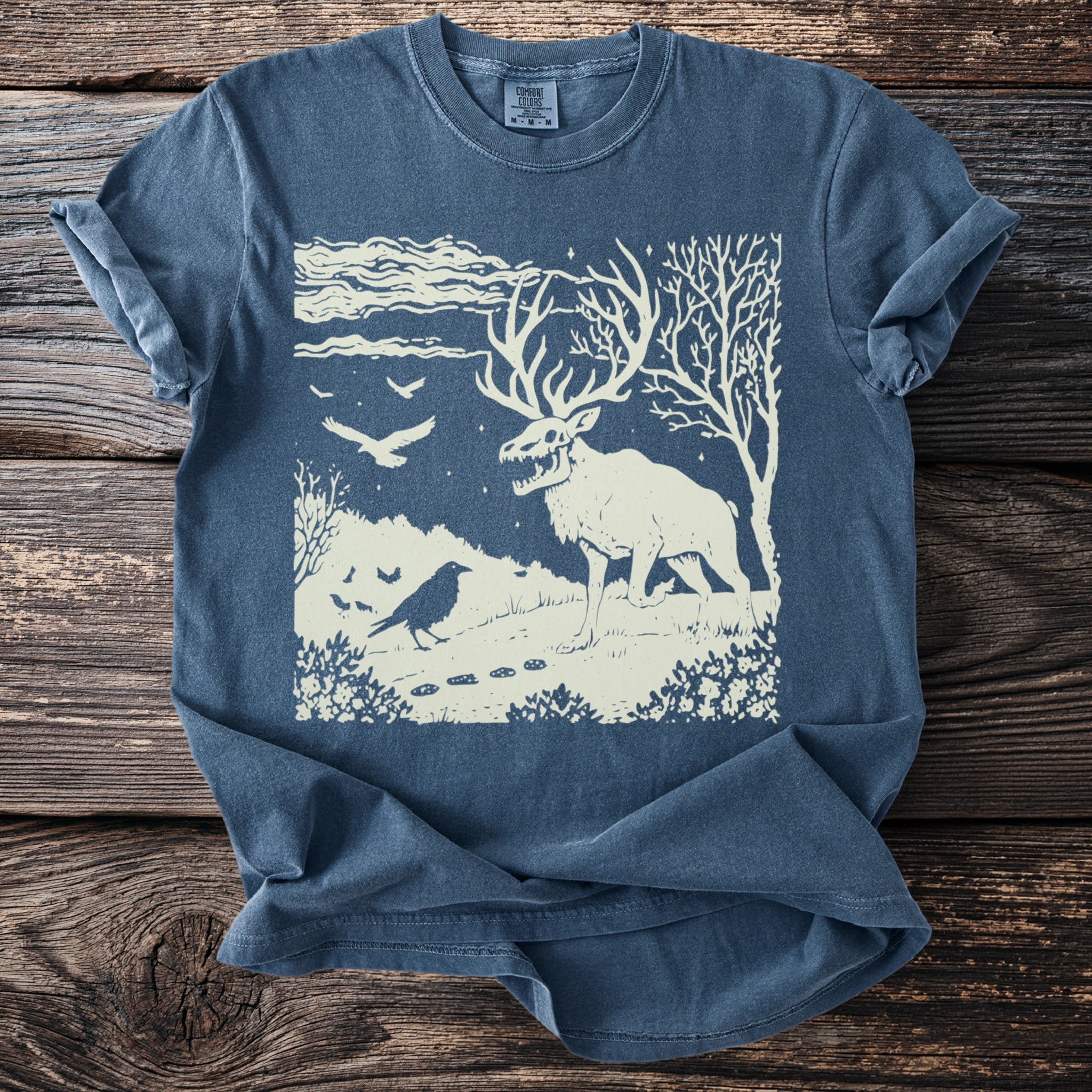 Skeletal stag with crow and birds on a starry night landscape graphic on a blue t-shirt.