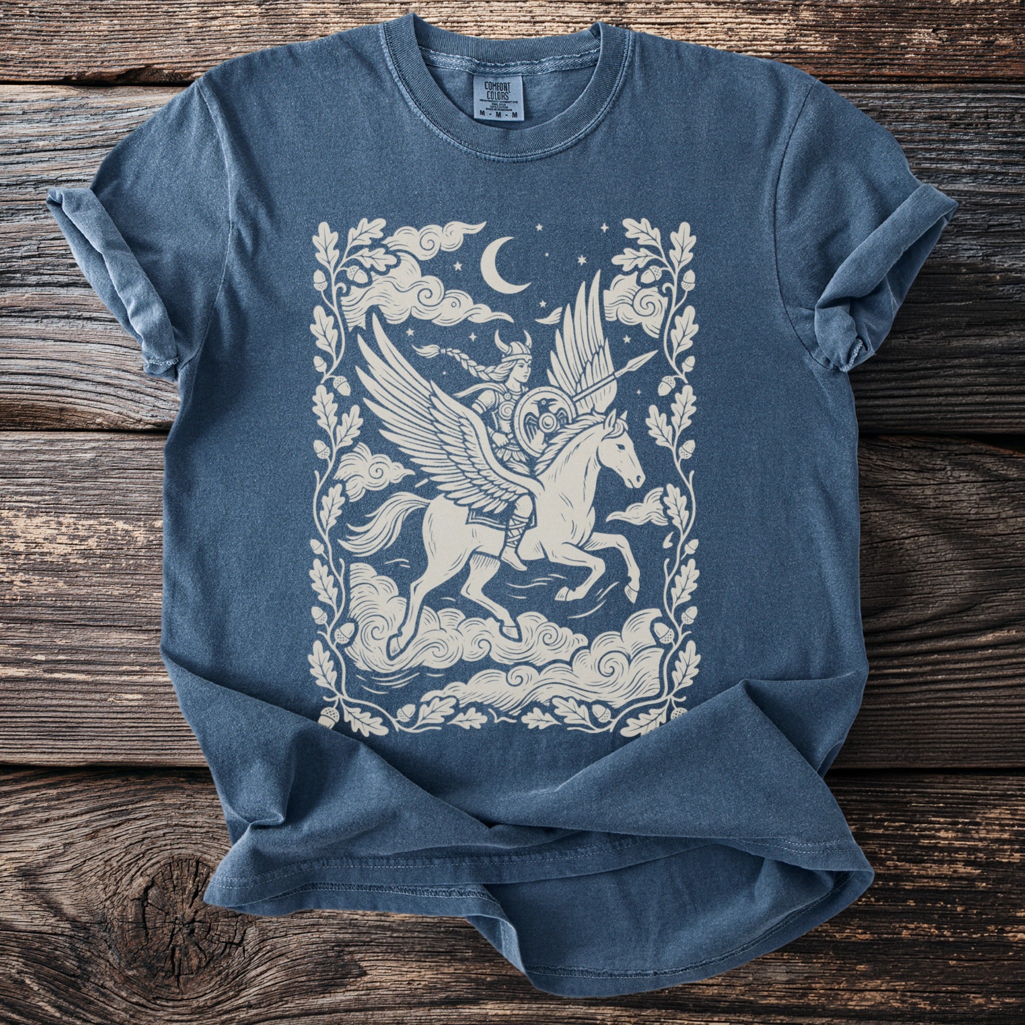 Blue t-shirt with cream illustration of a winged horse and rider in clouds under a moon.