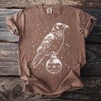 Brown t-shirt with a white graphic of a raven perched on a Christmas ornament with snowflakes.