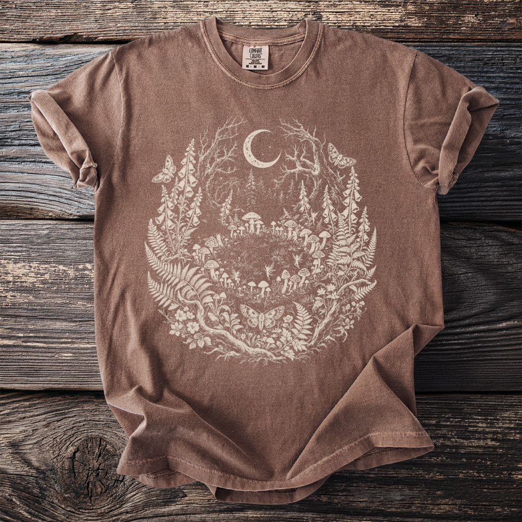 Brown t-shirt with a circular nature graphic featuring a moon, forest, mushrooms, fairies, and butterflies.