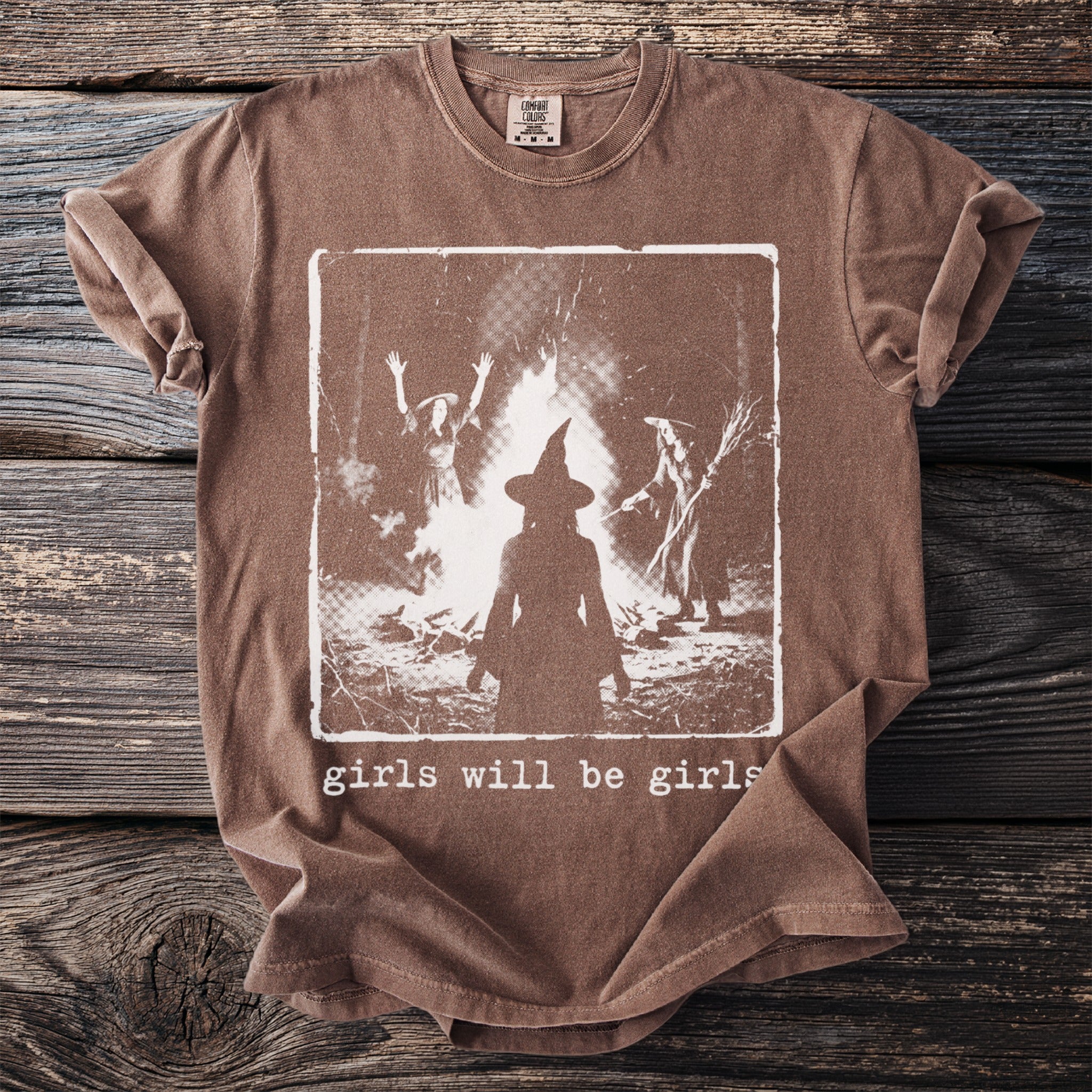 Brown t-shirt with graphic of three witches by a bonfire and the text "girls will be girls".