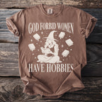 Brown t-shirt with "God Forbid Women Have Hobbies" text and witch reading book graphic.