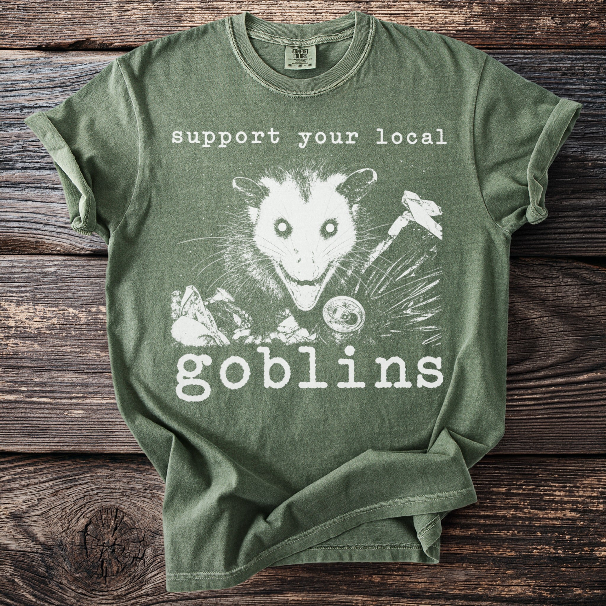 Green t-shirt with white graphic of an opossum, can, and tools, text says "support your local goblins".