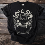 Black t-shirt with a cartoon illustration of a cat in a cauldron with moons and ornaments.