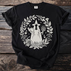 Black t-shirt featuring a white graphic of a witch holding a book and herbs surrounded by foliage.