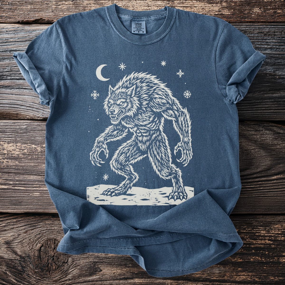 Denim blue t-shirt with a cream folk-cut print: a hunched werewolf, teeth bared and claws out, stands in the snow under a crescent moon.