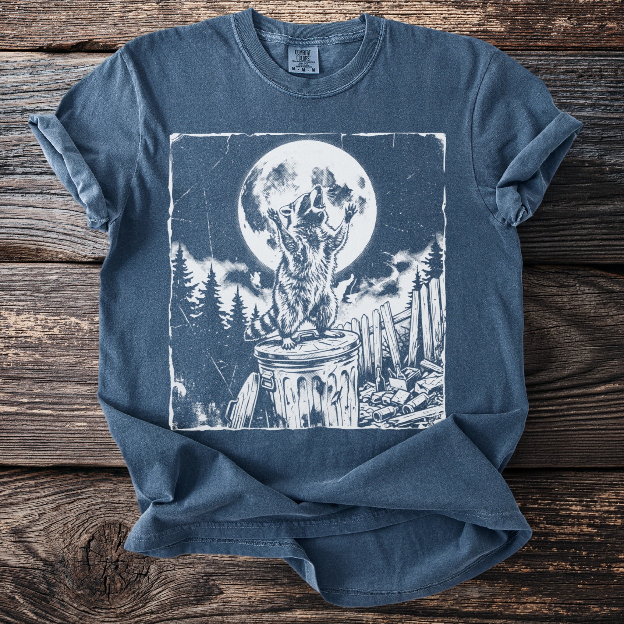 Blue t-shirt with a graphic of a raccoon standing on a trash can under a full moon.